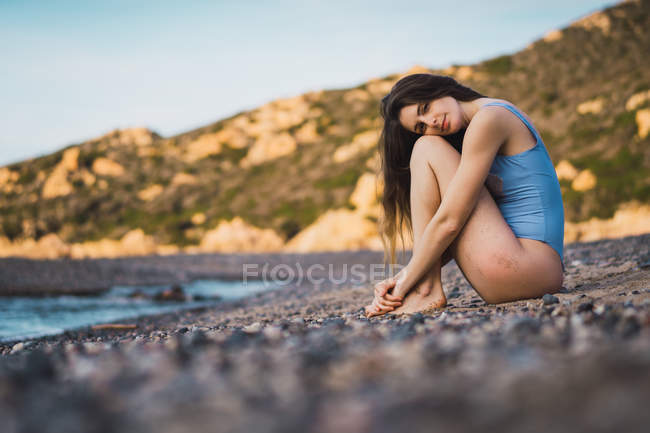 Giovane donna in costume da bagno seduta sulla spiaggia e guardando la fotocamera — Foto stock