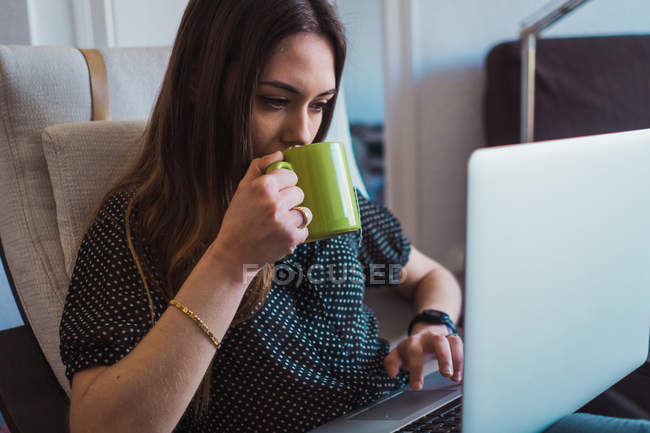 Femme utilisant un ordinateur portable dans un fauteuil et tenant une tasse de thé — Photo de stock