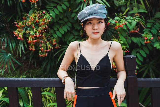 Jovem mulher asiática em roupas elegantes de pé em cerca no parque e olhando para a câmera — Fotografia de Stock