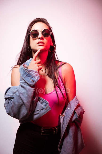 Seducir a la joven mujer vestida de rosa en gafas de sol de pie en la pared blanca - foto de stock