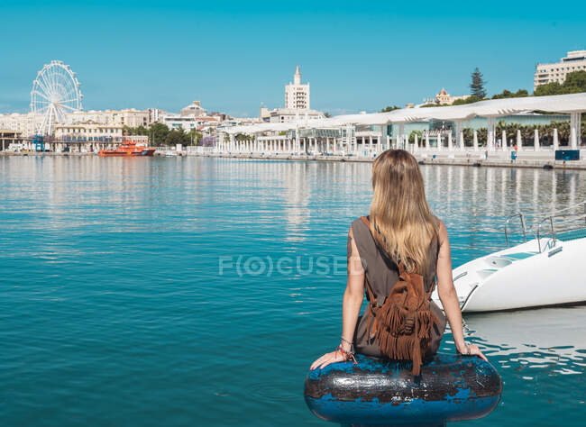 Visão traseira da mulher tatuada com pequena mochila sentada perto da água com as mãos afastadas e olhando para a zona costeira da cidade — Fotografia de Stock