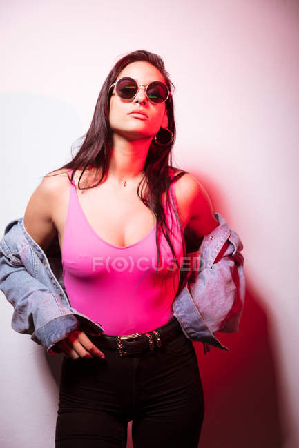 Jolie jeune femme rose habillée en lunettes de soleil debout au mur blanc — Photo de stock