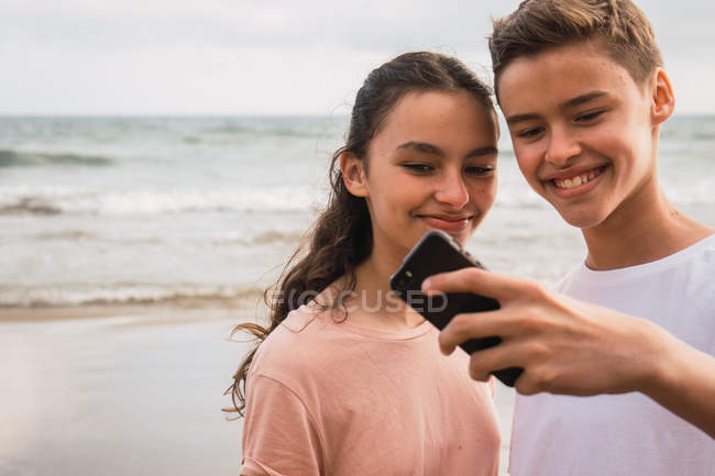 Sonriente adolescente y niña compartiendo teléfono inteligente en la orilla del mar - foto de stock