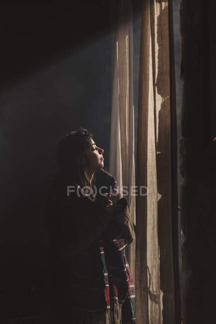 Вид збоку красива жінка, що стоїть з чашкою і насолоджується сонячним світлом з закритими очима біля вікна . — стокове фото