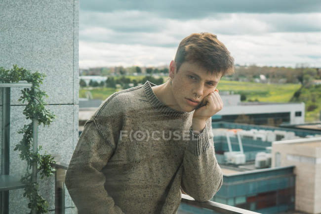 Вид через стекло серьезного молодого человека в свитере, опирающегося на руку на балкон и смотрящего в камеру — стоковое фото