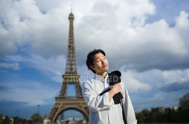 Pensativo chef japonés de pie con delantal frente a la Torre Eiffel en París - foto de stock