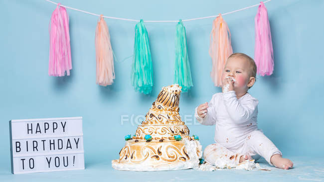 Cute little boy sitting at birthday cake and eating on blue background — Stock Photo