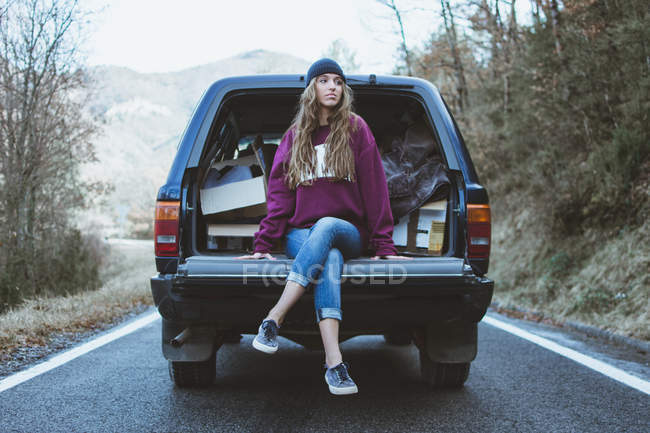 Nachdenkliche junge Frau sitzt auf Kofferraum von Auto im Wald — Stockfoto