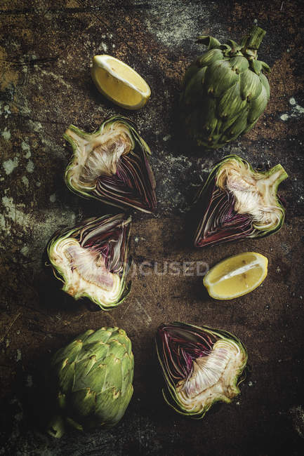 Alcachofas frescas cortadas a la mitad y trozos de limón sobre fondo oscuro oxidado - foto de stock