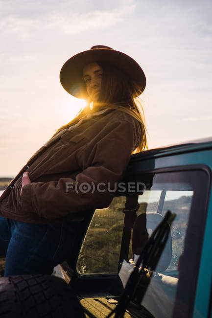 Portrait of woman in hat leaning on car at sunset — Stock Photo
