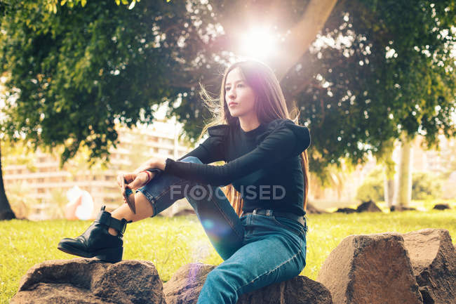 Jovem mulher pensativa sentada no rock com smartphone no parque — Fotografia de Stock