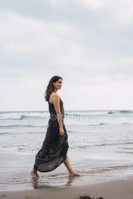 Элегантная женщина в черном платье прогулка по пляжу — стоковое фото