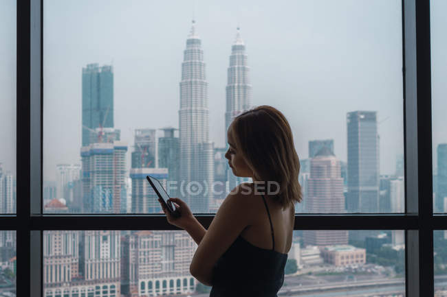 Schöne Frau mit Smartphone am Fenster in Wohnung mit Blick auf die Stadt — Stockfoto