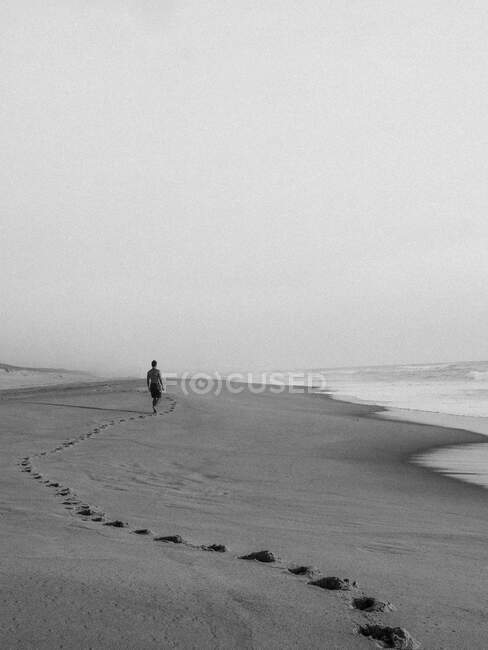 Plan noir et blanc d'un homme anonyme marchant sur un littoral sablonneux vide avec d'énormes vagues océaniques dans la brume — Photo de stock