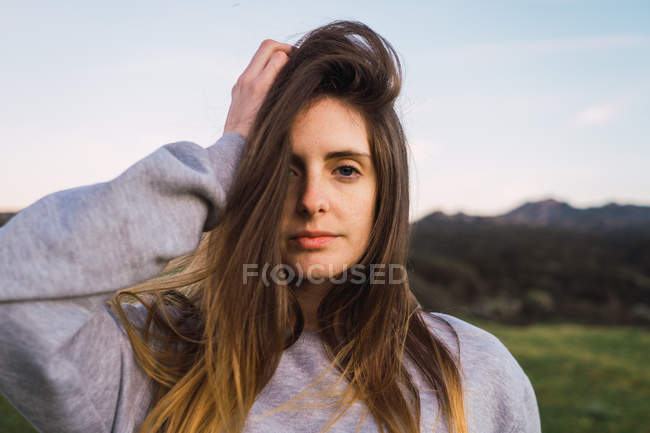 Mujer joven bonita mirando a la cámara y tocando la cabeza en la naturaleza - foto de stock