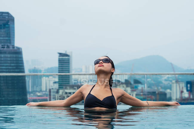 Азиатская женщина отдыхает в бассейне с современными небоскребами на заднем плане — стоковое фото
