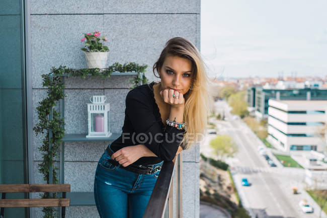 Blonde fille en tenue décontractée appuyé sur la clôture du balcon et regardant la caméra — Photo de stock