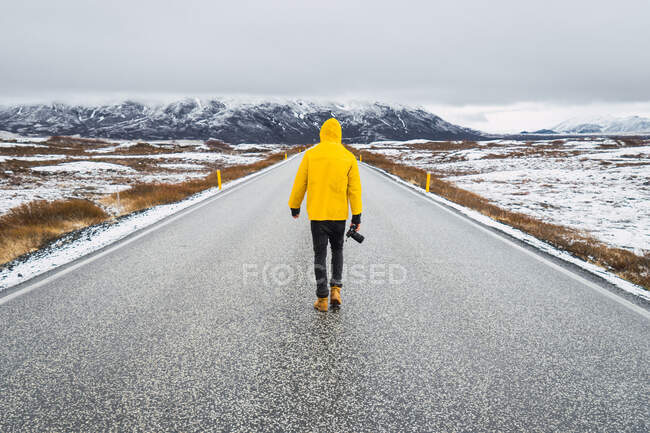 Back view of man in bright yellow coat holding photo camera and walking on long paved road in cold valley of Iceland — Stock Photo