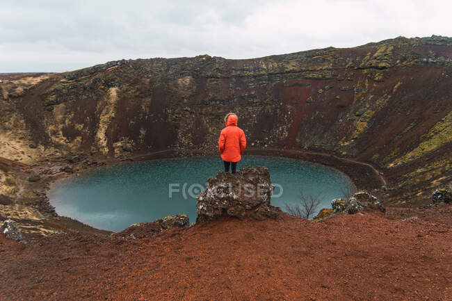 Back view of tourist in red coat standing on rock against small blue lake in basin of mountain in Iceland. - foto de stock