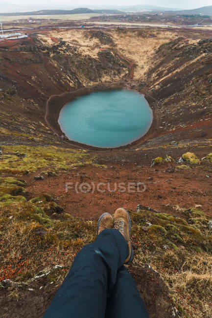 Beine eines Mannes, der an einer offenen Grube sitzt und auf einen kleinen See blickt, Island — Stockfoto