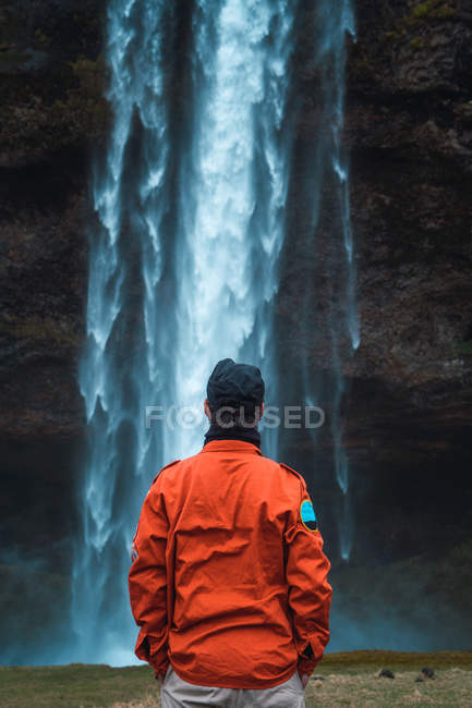 Hombre de abrigo de invierno naranja de pie con las manos en los bolsillos delante de la corriente de la cascada, Islandia - foto de stock