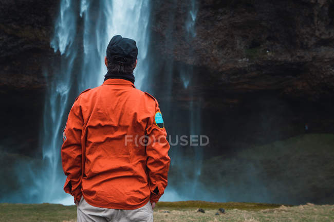 Mann in orangefarbenem Wintermantel steht mit Händen in Taschen vor Wasserfall, Island — Stockfoto