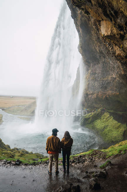 Casal de pé na colina sob o penhasco com cachoeira maravilhosa, Islândia — Fotografia de Stock