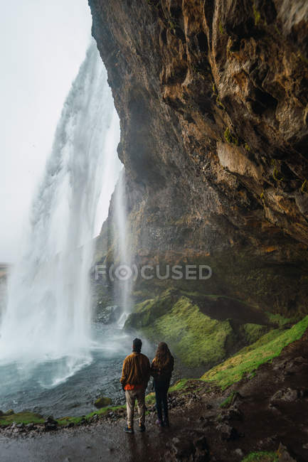 Casal de pé na colina sob o penhasco com cachoeira maravilhosa, Islândia — Fotografia de Stock