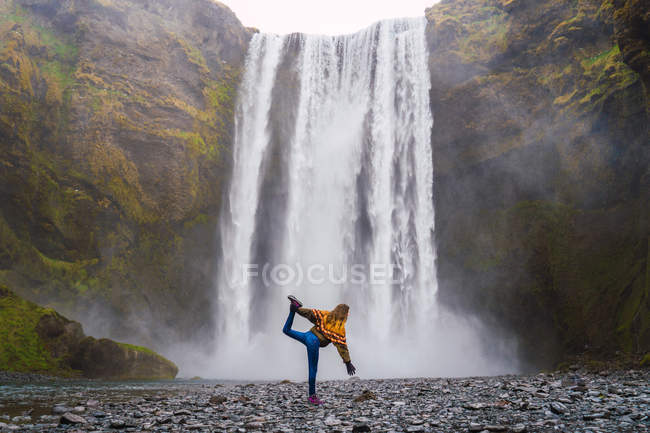 Женщина, стоящая в асане на гравии с водопадом на заднем плане, Исландия — стоковое фото