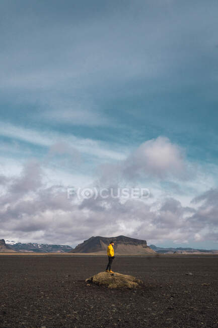 Homme anonyme debout sur un rocher au milieu d'une plaine rocheuse avec des montagnes derrière un ciel nuageux, Islande. — Photo de stock