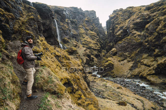 Bearded man with backpack and camera standing in mountains near small waterfall and river — Stock Photo