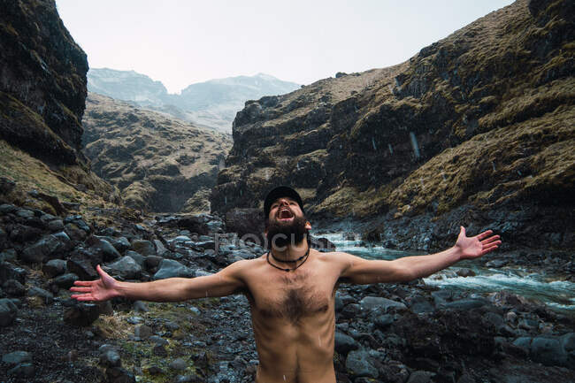 Young bearded shirtless male standing arms apart in mountains under rain shouting in air with joy — Stock Photo