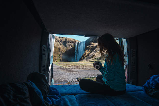 Woman sitting in cozy camping truck holding cup in front waterfall — Stock Photo