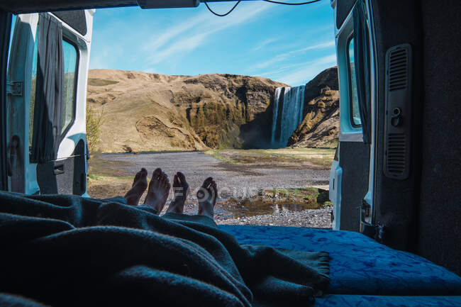Crop legs of male and female covered with blanket in camping truck near waterfall — Stock Photo