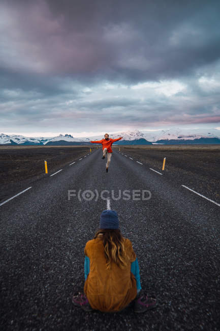 Mujer tomando foto de hombre bailando en camino vacío cerca de montañas nevadas - foto de stock