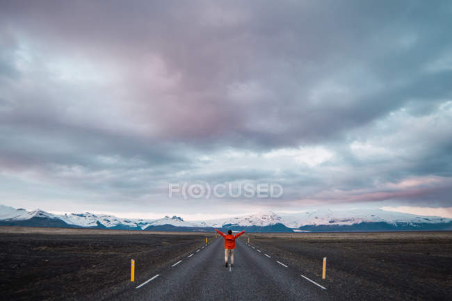 Homem de casaco vermelho andando na estrada com montanhas e céu dramático no fundo — Fotografia de Stock