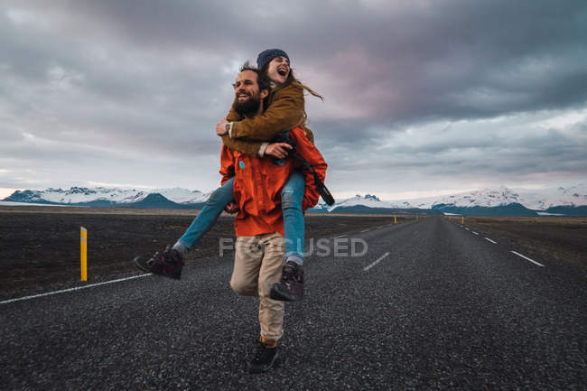 Barbudo hombre llevando mujer en espalda corriendo y riendo en el camino vacío cerca de las montañas - foto de stock
