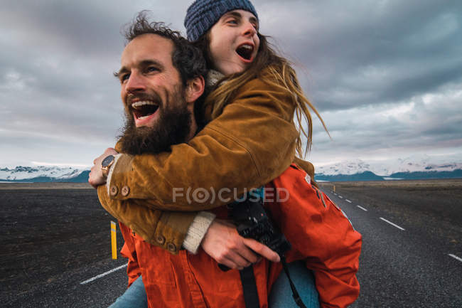 Barbudo hombre llevando mujer en espalda corriendo y riendo en el camino vacío cerca de las montañas - foto de stock