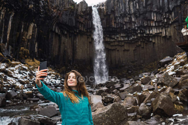Smiling young woman taking selfie waterfall and rocks with smartphone — Stock Photo