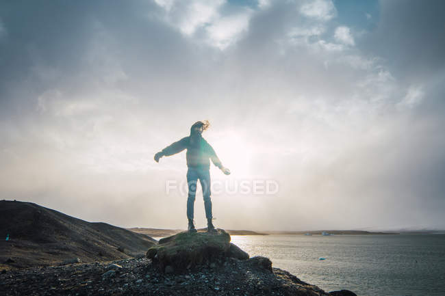 Женщина в капюшоне, стоящая на скале с видом на море и облака в солнечном свете — стоковое фото