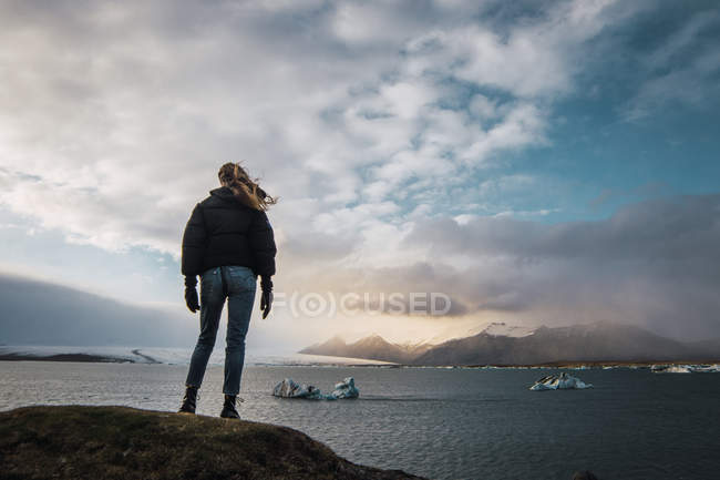 Turista de pie en el frío paisaje marino al atardecer y mirando a la vista - foto de stock