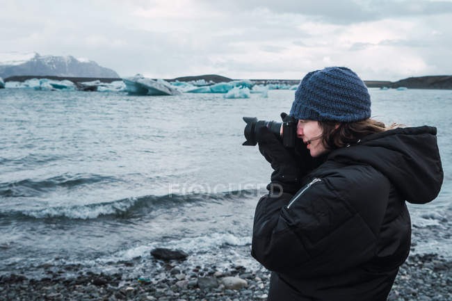 Femme en chapeau et vêtements chauds prenant des photos avec appareil photo de paysage marin de glace, Islande — Photo de stock