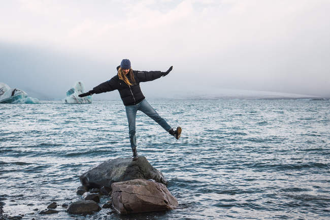 Mujer balanceándose sobre roca en agua fría azul del mar con trozos de hielo en el fondo, Islandia - foto de stock