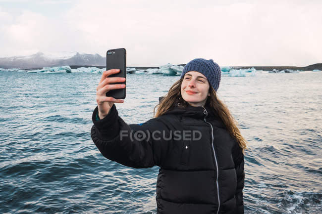 Sonriente joven mujer tomando selfie en frío playa - foto de stock