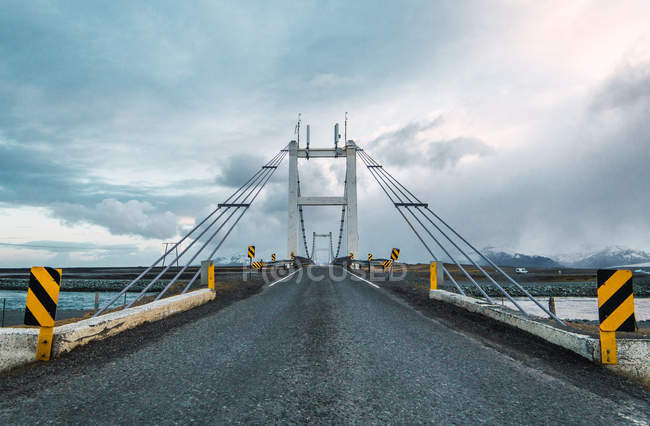 Paved road and suspension bridge under dark clouds, Iceland — Stock Photo