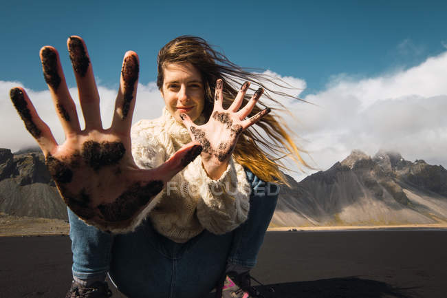 Mujer sonriente con el pelo ondulado mostrando las manos en arena negra con montañas en el fondo, Islandia - foto de stock