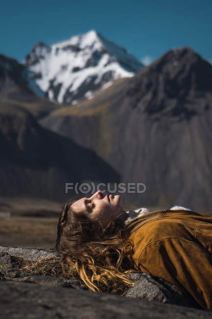 Mulher deitada com os olhos fechados na rocha com vista para as montanhas à luz do sol no fundo, Islândia — Fotografia de Stock