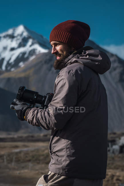 Бородатий чоловік тримає професійну камеру, стоячи на сонячному світлі гір — стокове фото