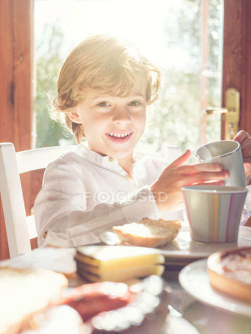 Niño pequeño con taza sentado a la mesa - foto de stock