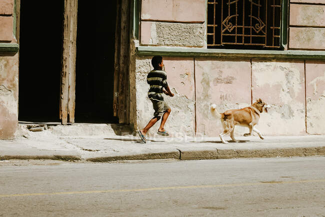 Kleiner Junge mit Hund läuft bei strahlendem Sonnenlicht über schäbiges Pflaster einer Straße in Kuba. — Stockfoto